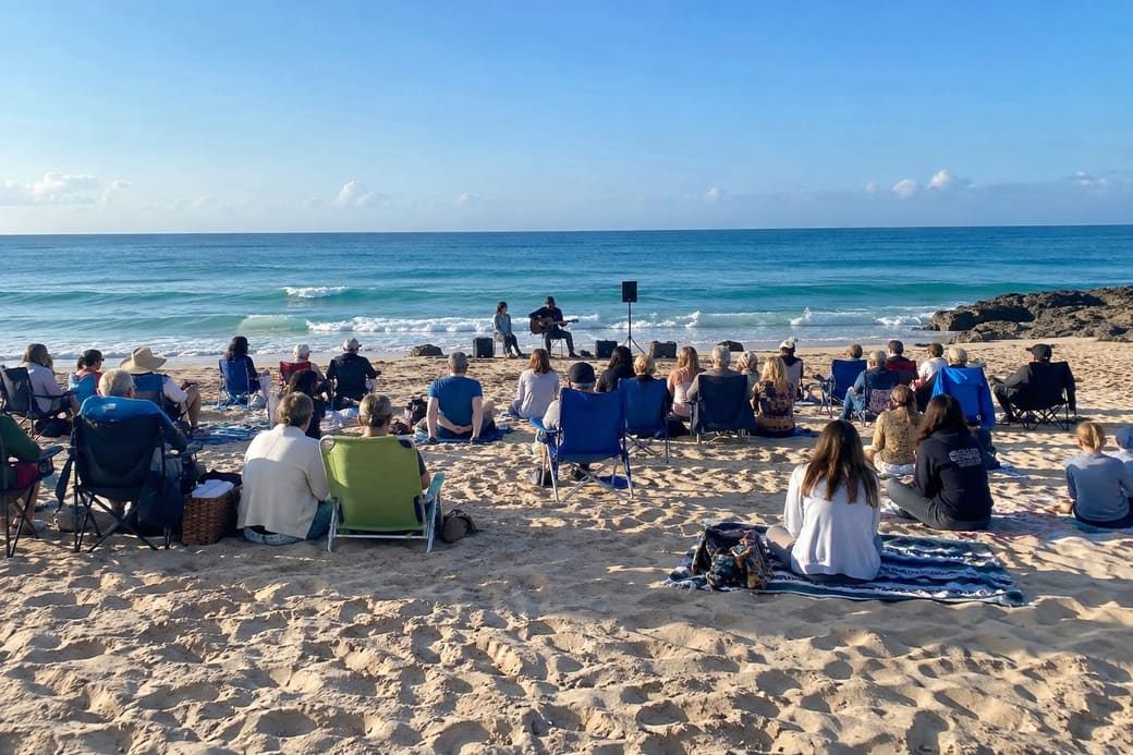 People on folding chairs and blankets on the sand, gathered for a Sunday morning service by the water